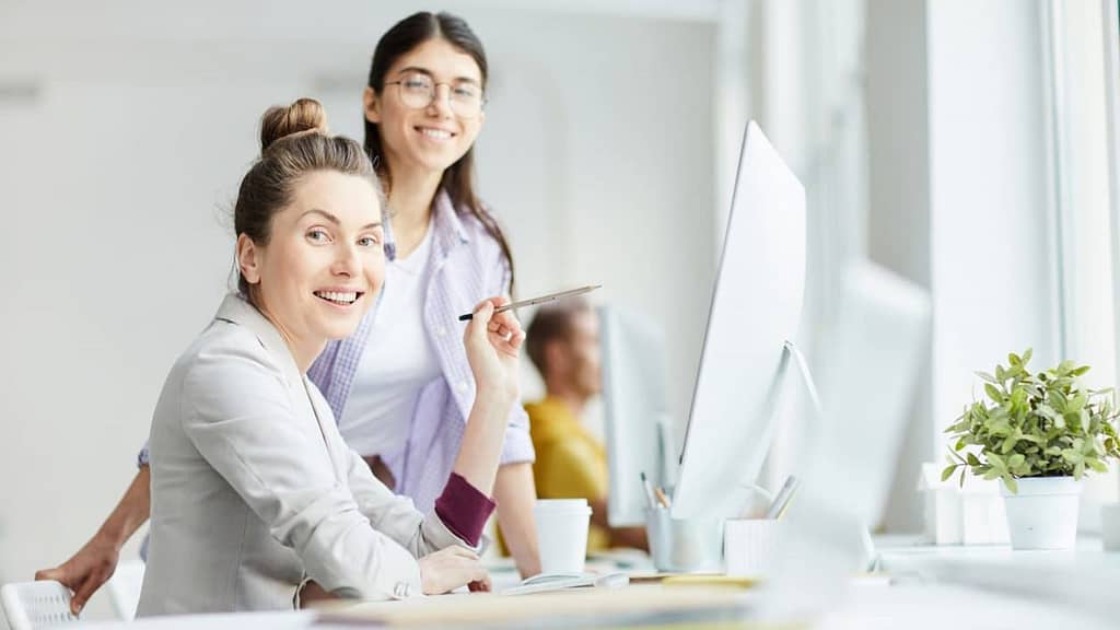 two career women smiling at work with white monitors and plants